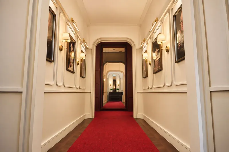 A cream corridor with framed portraits, wall lamps and a red carpet leading inside