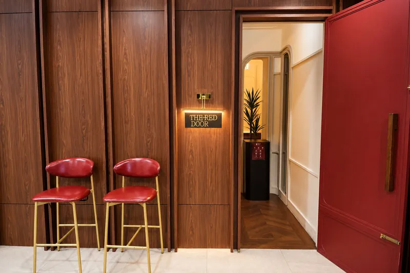 The red door of the venue open onto a cream corridor, two red stools by a wooden wall