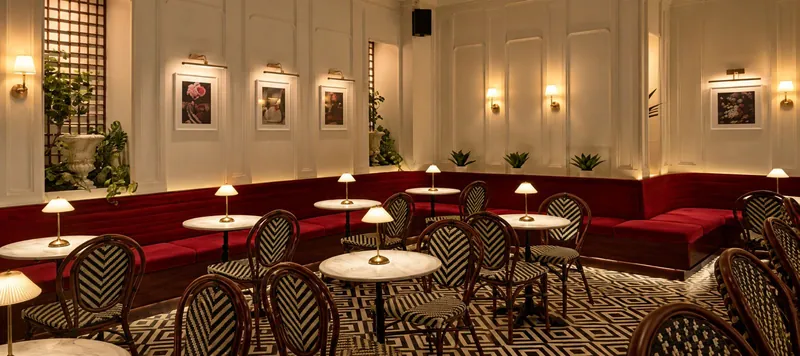 A white salon with red velvet banquettes, marble tables, bistro chairs and framed flower prints