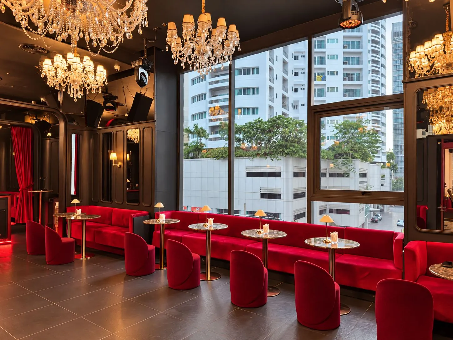 A lounge by tall windows with red velvet seats and chandeliers, city view outside